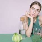A young woman in a green top sitting with vegetables and a glass of water.